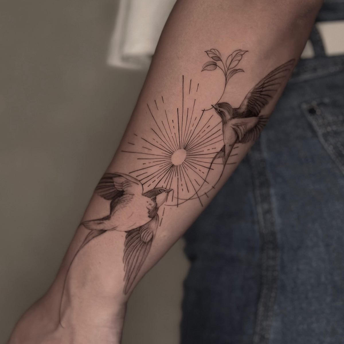 Fine-line swallows flying around sun rays, tattooed on a forearm